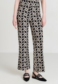 Femme portant un pantalon en motif géométrique beige et noir avec des escarpins à bout pointu et bride arrière noirs ornés de clous argentés et de boucles.