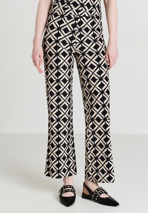 Frau trägt beige und schwarze geometrisch gemusterte Hosen mit schwarzen Slingback-Schuhen mit spitzen Zehen, die mit silbernen Nieten und Schnallen verziert sind.