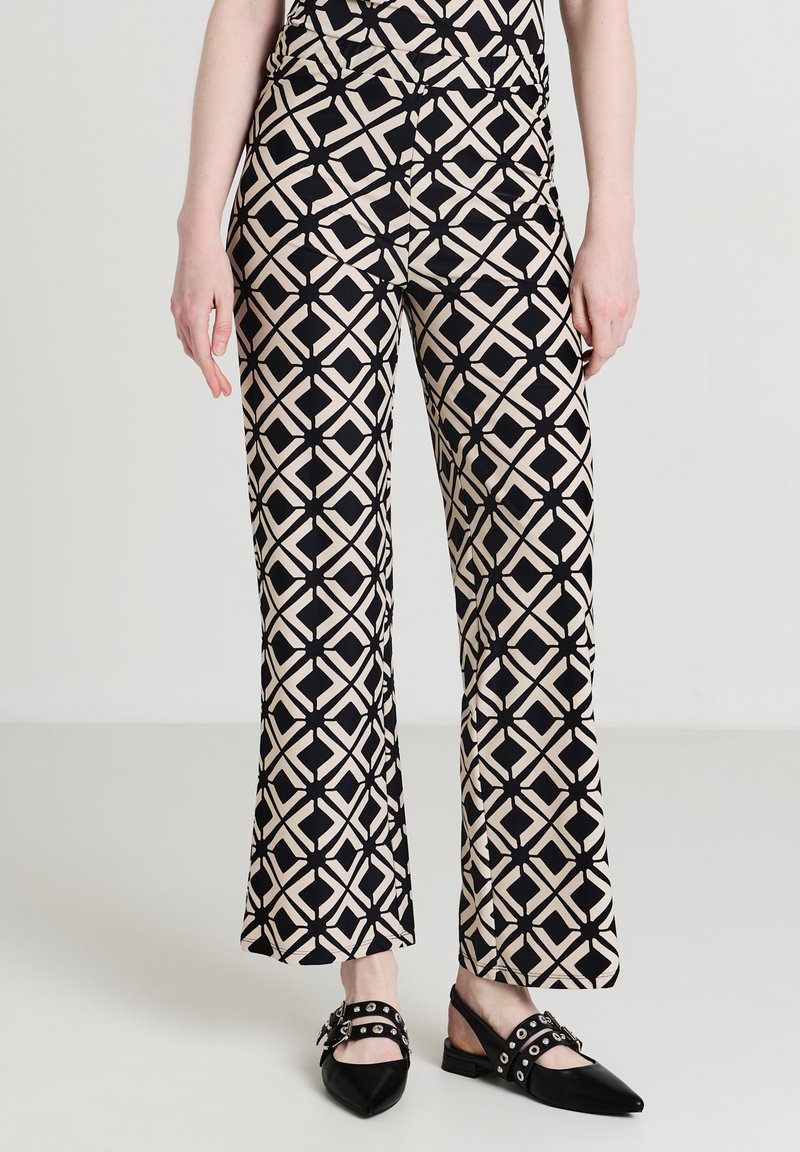 Femme portant un pantalon en motif géométrique beige et noir avec des escarpins à bout pointu et bride arrière noirs ornés de clous argentés et de boucles.