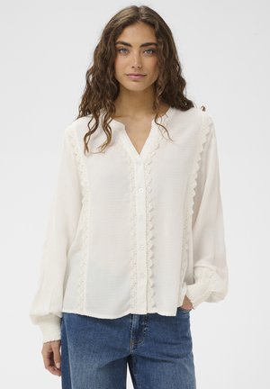 Valge, tekstuuriga viimistlusega pika varrukaga pluus, nööpide ääres pitsi detailid ja elastsete mansettidega, kombineeritud siniste teksapükstega.