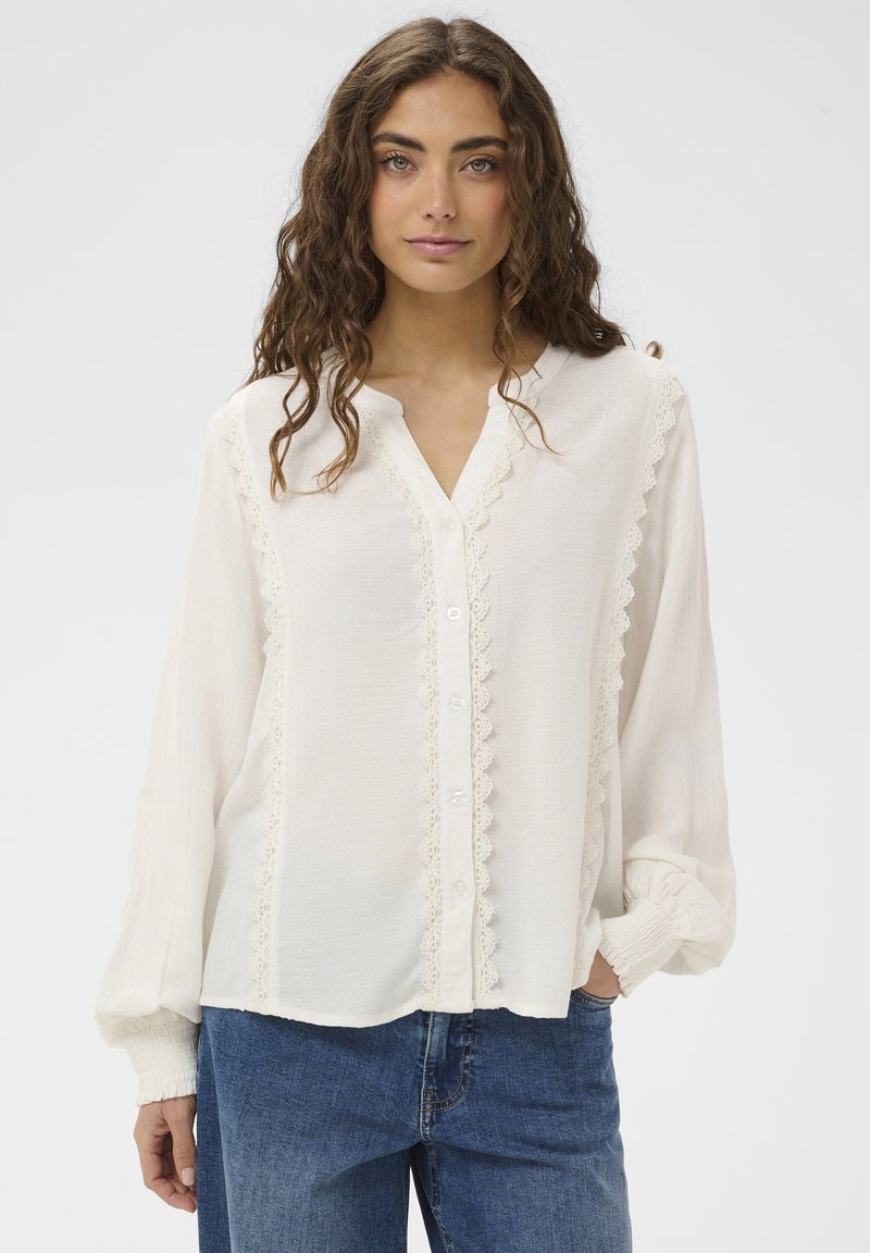 Langärmlige weiße Bluse mit strukturierter Oberfläche, Spitzenakzenten entlang der Knöpfe und elastischen Bündchen, kombiniert mit blauen Jeans.