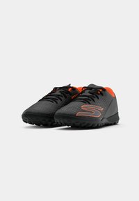 Zapatillas deportivas negras con detalles en naranja, que cuentan con material texturizado, puntera redondeada y suela rugosa para mayor tracción.