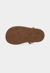 Καφέ σόλα σανδάλι με ανάγλυφα σχήματα εμπνευσμένα από τη θάλασσα, όπως αστερίες, κοχύλια και σπείρες, και λογότυπο "POM D'API".