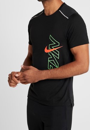 Mężczyzna w czarnej koszulce Nike z krótkim rękawem z neonowo zielonym i pomarańczowym logo, stoi ze splecionymi rękami na jednolitym tle.