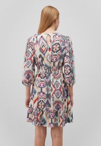Blumenmuster-Kleid mit Puffärmeln, geraffter Taille und Schlüsselaugen-Rückendetails. Die Farben umfassen Lila, Rot und Blau auf einem cremefarbenen Hintergrund.