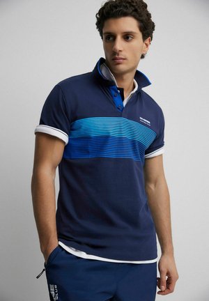 Młody mężczyzna w granatowej koszulce polo z niebieskimi poziomymi paskami i białym wykończeniem, patrzy w bok z rękami w kieszeniach granatowych spodni.