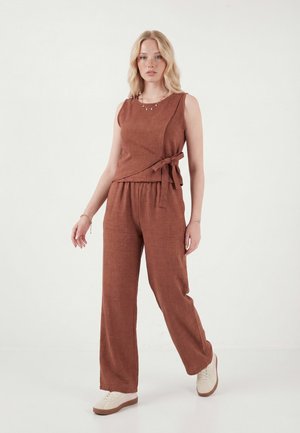 Nő barna ujjatlan átkötős felsőben és hozzáillő bőszárú nadrágban, fehér tornacipővel, sima világos háttér előtt állva.