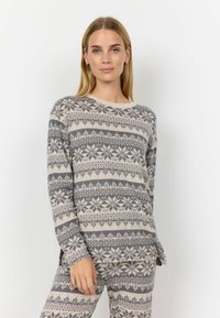 Frau in passendem beigen und grauen gemusterten Langarmoberteil und Hose mit Schneeflocken- und geometrischen Designs, stehend vor einem schlichten Hintergrund.