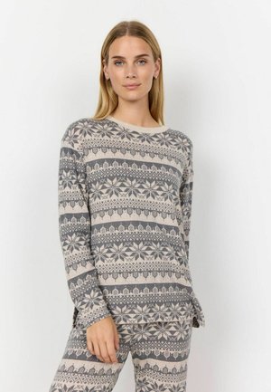 Kvinna som bär en matchande mönstrad långärmad topp och byxor i beige och grått med snöflinge- och geometriska mönster, stående mot en enfärgad bakgrund.