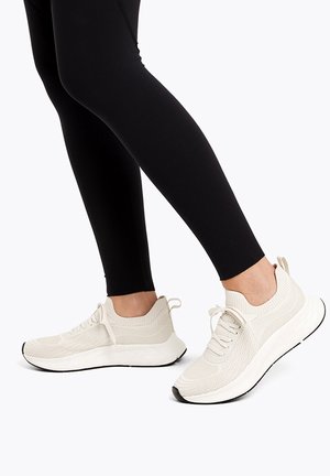 Person iført hvide strikkede sneakers og sorte leggings, stående med den ene fod let hævet på en hvid baggrund.