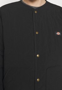 Black quilted jacket with brass snap buttons, two front pockets, and a small rectangular Dickies logo patch on the chest.