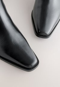 Bottes noires en cuir synthétique à la cheville avec un bout pointu et une finition texturée. Talon carré et semelle légèrement crantée.