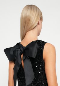 Abito nero in paillettes con un grande fiocco in satin nero sul retro. La trama è liscia, con decorazioni in paillettes lucide e un design con scollatura.