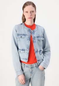 Ανοιχτό μπλε τζιν μπουφάν με στρογγυλή λαιμόκοψη, μπροστινές τσέπες, ασημί κουμπιά και διακριτές ραφές, φορεμένο πάνω από ένα κόκκινο πουκάμισο.