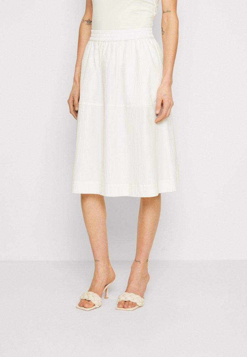 Femme portant une jupe midi blanche à taille élastique et des sandales à talons tressées crème, debout devant un fond uni.