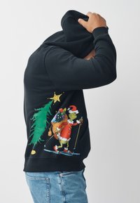 Zwarte hoodie met een kleurrijke afbeelding van de Grinch die aan het skiën is, gekleed in rood met witte accenten, omringd door kerstbomen en een ster.