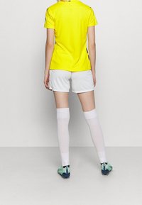 Gelbes Sporttrikot, weiße Shorts und knielange weiße Socken. Ausgestattet mit grünen und blauen Fußballschuhen. Sauberes, körperbetontes Design.