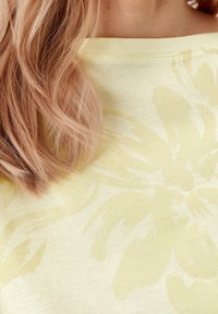 Žlutá látka s květinovým vzorem a jemnou strukturou, s širokým výstřihem. Světlé vlasy částečně viditelné po straně.