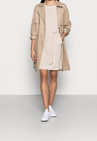Manteau beige sur une robe légère à rayures avec une ceinture à nouer, associé à des sneakers blanches. Tissu lisse, design mi-long et coupe décontractée.