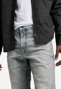 Svart jacka över en vit skjorta, tillsammans med ljusblå, smalare jeans med en subtil blekning och en liten logotagg på fickan.