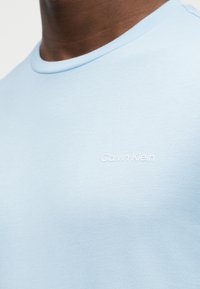 Κοντινό πλάνο ενός ατόμου που φοράει ανοιχτό μπλε μπλουζάκι με στρογγυλή λαιμόκοψη και μικρό λευκό λογότυπο "Calvin Klein" στο στήθος.