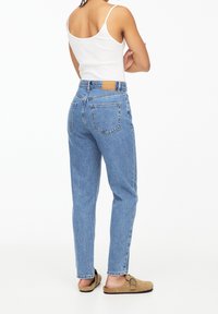 Persona con jeans azules de tiro alto, camiseta blanca sin mangas y zapatos de tela color beige, de pie con los brazos cruzados y de espaldas sobre un fondo blanco.