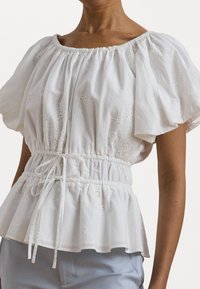 Vit bomullskjorta med puffärmar, rynkad halsringning och cinchad midja. Har blommig broderi och en flödande peplumdesign.