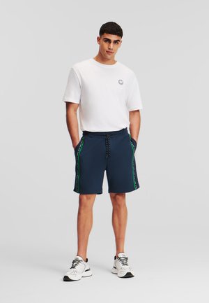 Marineblauwe shorts met groene zijstrepen, elastische tailleband en trekkoord, gecombineerd met een witte T-shirt met ronde hals. Model staat met de handen in de zakken.