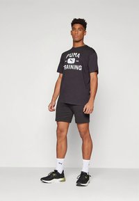 Svart bomulls T-skjorte med "PUMA TRAINING" grafikk, kombinert med mørke shorts og svarte joggesko. Modellen står oppreist med et nøytralt uttrykk.