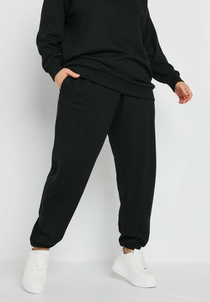 Personne portant un pantalon de survêtement noir et un sweat-shirt assorti, debout avec une main dans la poche, des baskets blanches, sur un fond clair uni.