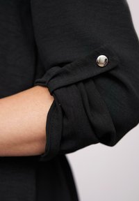 Opgerolde zwarte lange mouw vastgezet met een zilveren drukknoop op een lichtgekleurde arm tegen een effen achtergrond.