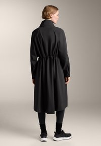 Zwarte, lange mouwjas met een getailleerde taille, gemaakt van soepel materiaal. Heeft een hoge kraag en valt tot halverwege de kuit, gecombineerd met donkere leggings en schoenen.