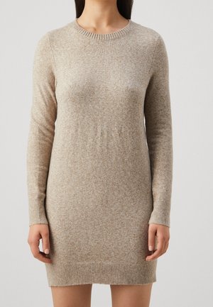 Femme portant une robe-pull en maille beige à manches longues avec poignets et ourlet côtelés, sur un fond gris uni.