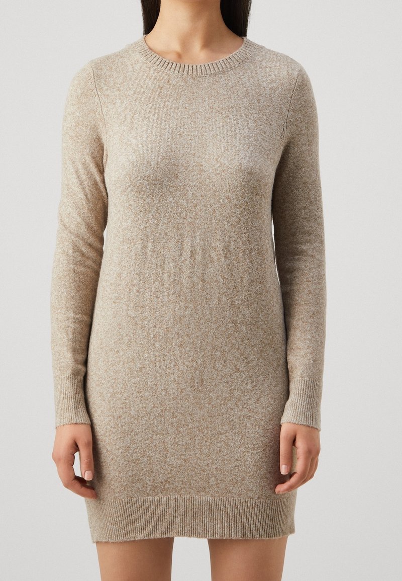 Femme portant une robe-pull en maille beige à manches longues avec poignets et ourlet côtelés, sur un fond gris uni.