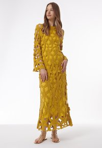Robe au crochet jaune à manches longues, présentant des motifs floraux complexes et des découpes. L'ourlet est légèrement évasé, assorti à des sandales dorées.