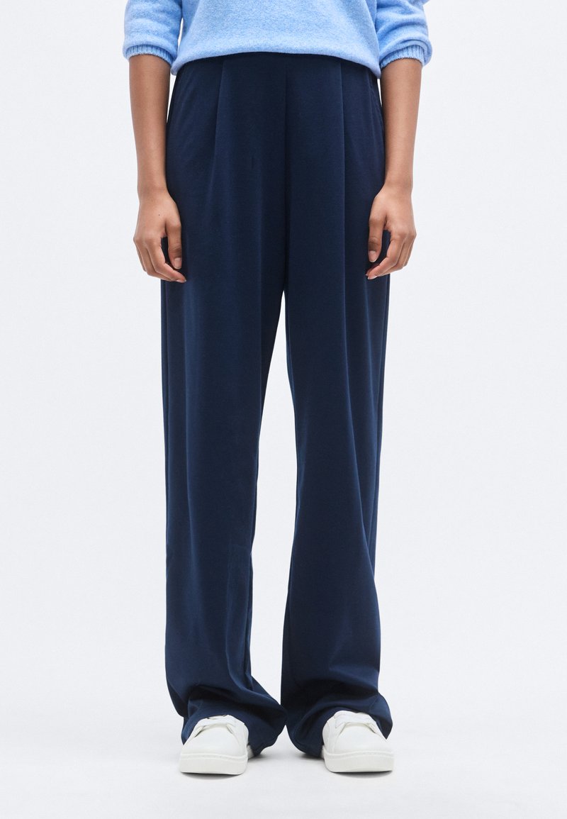 Φαρδιά παντελόνια σε ναυτικό μπλε από μαλακό ύφασμα, με πιέτες, ελαστική μέση και συνδυασμένα με λευκά αθλητικά παπούτσια.
