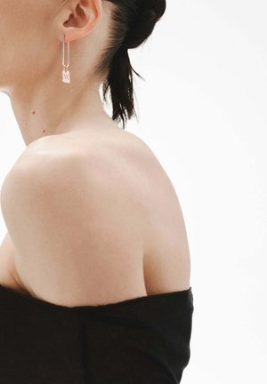 Los pendientes de aro de plata se sujetan a charms rectangulares. Los pendientes tienen un acabado pulido y se llevan con una blusa sin hombros sobre un fondo blanco.