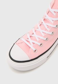 Pink canvas sneaker with white laces and silver eyelets, featuring a black rubber sole and white toe cap. Textured fabric and stitching details visible.