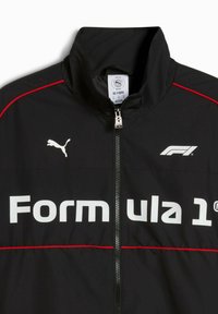 Sort vindjakke med røde detaljer og høj krave. Har store hvide "Formula 1" bogstaver og et hvidt Puma-logo på brystet.