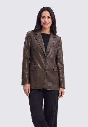 Vrouw met lang donker haar draagt een donkerbruine leren blazer over een zwart topje en zwarte broek, staand tegen een effen lichte achtergrond.