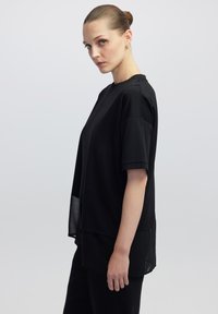 Schwarzes, übergroßes T-Shirt mit kurzen Ärmeln, ausgestattet mit einem Rundhalsausschnitt, einem schichtartigen Design und einem leicht transparenten Saum. Glatte Stofftextur.