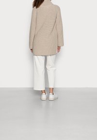 Beżowy sweter z wysokim kołnierzem, długimi rękawami i luźnym fasonem, w połączeniu z białymi spodniami 3/4 i białymi sneakersami. Gładkie tło.