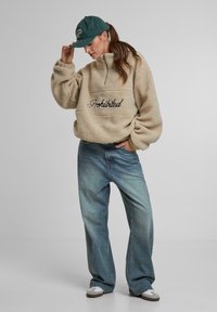 Pullover in pile crema con testo "Proibito", design a mezza zip. Abbinato a jeans larghi blu chiaro e un cappellino verde scuro, con logo.
