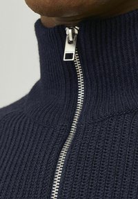 Navyblauer Strickpullover mit erhabener Rippstruktur, ausgestattet mit einem hohen Kragen und einem silberfarbenen Reißverschluss, der elegante Hardware-Details betont.