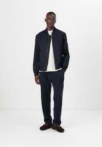Veste bombardier navy à fermeture éclair à l'avant, poignets côtelés et poche latérale ; portée sur une chemise blanche et associée à un pantalon navy assorti et des chaussures marron.