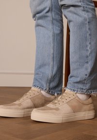 Beige Wildlederschuhe mit strukturierten Akzenten und einer Gummisohle, kombiniert mit hellblauen, ausgefransten Jeans. Holzstuhl im Hintergrund.