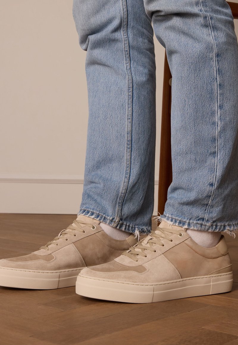 Beige Wildlederschuhe mit strukturierten Akzenten und einer Gummisohle, kombiniert mit hellblauen, ausgefransten Jeans. Holzstuhl im Hintergrund.
