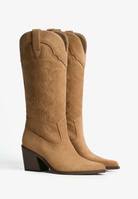 Tan mockaskängor i cowboystil med västerländsk sömnad, spetsiga tår och klädd klack. Designen har en vågig topp och dragremmar.