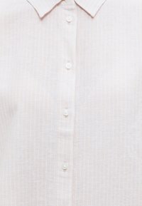 Ανοιχτό μπεζ πουκάμισο με γιακά, με διακριτικές κάθετες ρίγες και λευκά κουμπιά στο μπροστινό μέρος κατά μήκος.