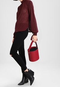 Burgundy teksturoitu paita, jossa on kuplamaiset hihat, yhdistetty mustiin revittyihin skinny-farkkuihin ja nilkkureihin, pitäen pyöreää punaista ämpärikassia.